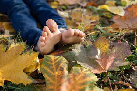 barefoot child sits on the ground among fallen yellow autumn leaves in park. cute children's toes...