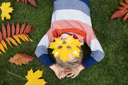 Hello Autumn. seasonal atmospheric photo. love autumn and interesting ideas for creativity. the child lies on the grass and hides his face behind an autumn maple leaf on which a cat's face is carved.の写真素材