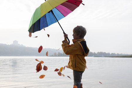 On a cloudy day, a child stands under a rainbow umbrella from which autumn leaves are falling. autumn atmosphere. Seasonal joys, cheerful childhood. Hello Autumnの写真素材