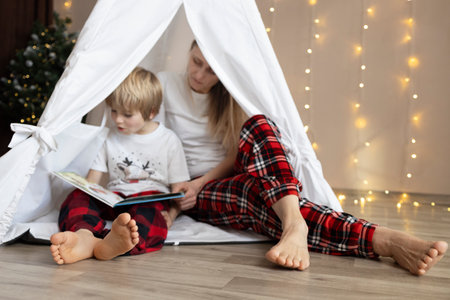 barefoot mother and son in pajamas sit in a play tent in the room and read book. Cozy atmosphere. Christmas Eve with family. concept of spending quality time together at home. Selective focus on feetの写真素材