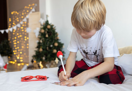 little boy writes a letter to Santa Claus and dreams of a gift against the backdrop of decorations and garland lights indoors. Merry Christmas and happy holidays. A wish list.の写真素材