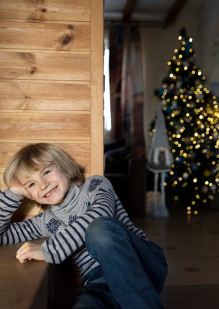 joyful boy in a sweater is sitting in a room at home, with Christmas tree lights behind him. Happy Christmas waiting concept. cozy pictures on a New Year's theme. positive childの写真素材