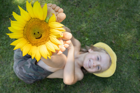 cheerful child lies on the grass, on his bare feet there is a sunflower with a carved smile. Focus on big flower. Summer mood, floral fantasies, atmosphere of positivity and fun. Creative farming, agricultureの写真素材
