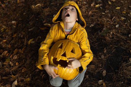 frightened child in an orange jacket sits on the ground strewn with fallen leaves, holding a large Halloween pumpkin in his hands. Evening forest. mood to scare and have fun. Trick or treatの写真素材