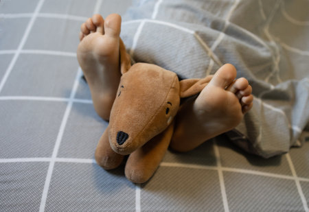 close-up of child's bare feet on the bed under a blanket, a soft funny toy dog lies on them. Good morning. Good night. A sleeping cozy place, sleep with pleasure in the company of your favorite toyの写真素材