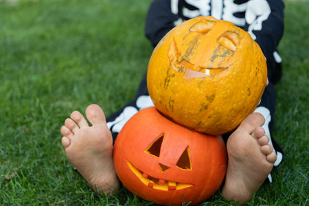 bare feet of a child sitting on the grass, with carved orange pumpkins with a Halloween grin. Preparing for Halloween. cheerful childhood, pampering. Don't be afraid, have funの写真素材