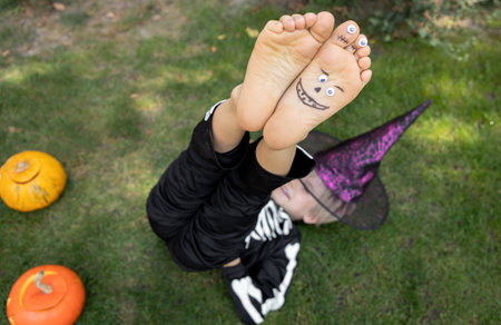 barefoot child in a skeleton costume plays around lying on the grass with his legs raised up. Halloween concept. Children's creativity, positive fun moments. holiday joys Selective focusの写真素材