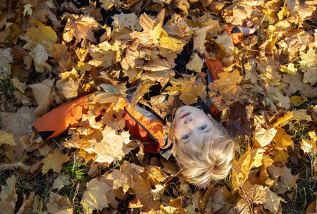 child lies under a lot of bright fallen leaves in a park or forest. Boy having fun outdoors. Enjoying autumn. Hello Autumn. Seasonal walksの写真素材