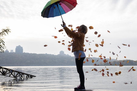 On a cloudy day, a woman has fun standing under a rainbow umbrella from which autumn leaves are...