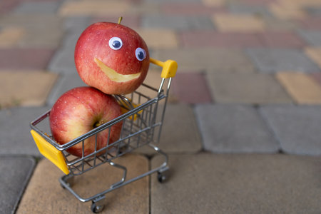 small toy grocery cart with two delicious red apples, one of which has a smile cut out and eyes glued on. Funny advertisement for organic products in a supermarketの写真素材