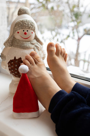 toy snowman, Santa hat, bare feet of a child sitting on the windowsill by the window. Waiting for a Christmas miracle. Cozy, festive atmosphere. Santa's Helperの写真素材