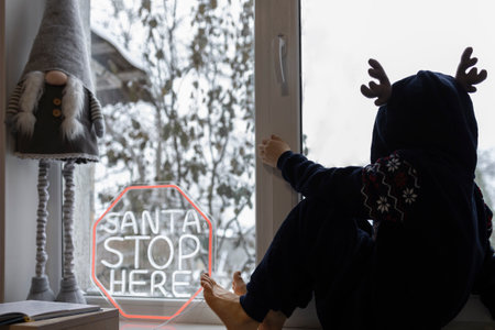 child in a plush deer costume sits on the windowsill near the window and waits for Santa. Waiting for the Christmas holidaysの写真素材
