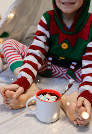 cute child dressed as a Christmas elf sits barefoot on the floor and drinks cocoa with marshmallows. Cozy homely holiday atmosphereの写真素材
