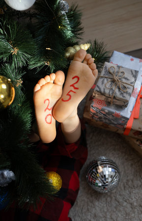Christmas and New Year celebration concept. a barefoot child lies on the floor near the Christmas tree. The numbers 2025 are painted on the legs. Waiting for gifts.の写真素材