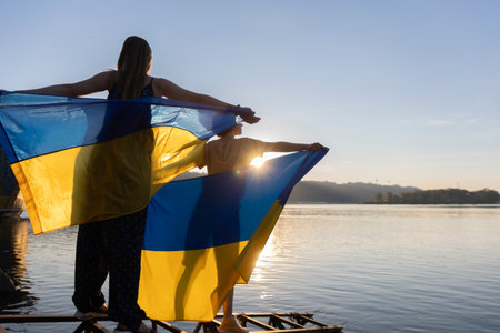 two women hold Ukrainian yellow-blue flags behind back, silhouetted against of sunset sky and riveの写真素材