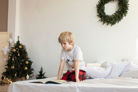 6-7 year old boy in pajamas sits on the bed and reads a book. Christmas Eve at home. Waiting for the holidays. Hobby reading booksの写真素材