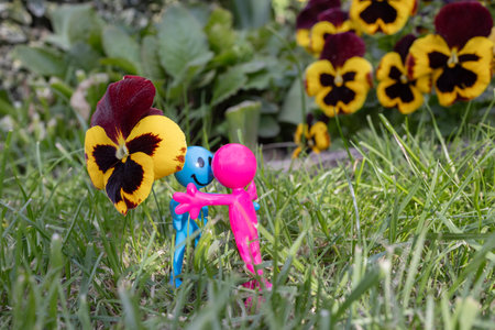 funny souvenir figurines are happy to meet each other, standing in the grass near large pansy flowers. couple in love. concept of romance, love and tender feelings, romantic surpriseの写真素材