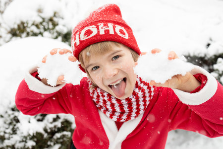 face of cute cheerful boy 7 years old in striped red scarf and Santa hat with inscription hohoho. Winter walks, snowy weather. happy childhood, winter holidays, stick out your tongue and taste snowの写真素材