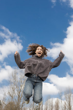 Cheerful adult teenage girl with curly hair in casual clothes jumps up enthusiastically with her arms raised. Emotion, surprise, anticipation. Adolescence. Body energy, outdoor activitiesの写真素材
