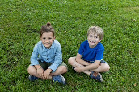 Two happy boys are sitting on the grass with their legs crossed. summer rest in the park or in the school yard. positive, joy. having fun together. friends forever. Summer mood, carefree joyful childhoodの写真素材