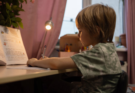 A 6-year-old boy sits at the table and carefully writes his homework. Schoolboy doing homework in the room, turning on the desk lamp. self-studyの写真素材