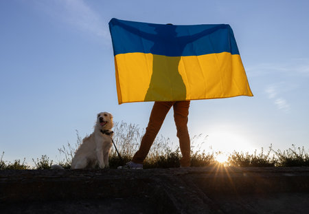 woman holds large flag behind her back, dog sits next to her against backdrop of the sunset sky. An evening walk with a beloved pet.の写真素材