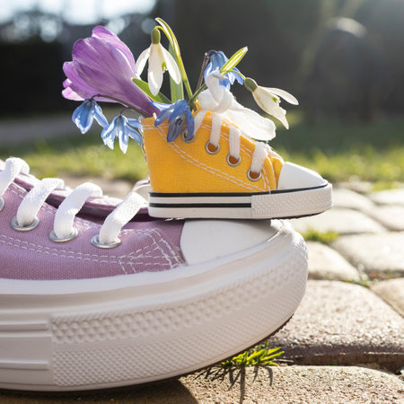 A small yellow sneaker with spring flowers stands on a big adult sneaker on a sunny day. Hello spring. Festive cute card. Be positive, have a nice dayの写真素材