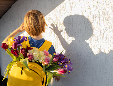boy with bright yellow backpack on his back full of colorful spring flowers, tulips. irises, roses. Interesting shadow from child on wall. Little courier, a gift with love for mom. Mother's Dayの写真素材