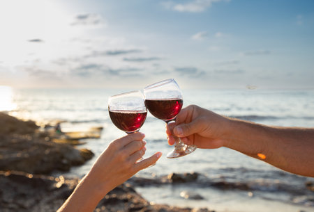 Glass goblets with red wine in male and female hands against the sky on the sea coast. Romantic evening for twoの写真素材