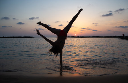 silhouette of slender girl doing one-arm upside down flip on seashore against backdrop of a sunset. concept of cheerfulness, joy, happiness, healthy lifestyle. be active and positive. hello holidaysの写真素材