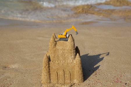 miniature orange toy excavator stands on a sand castle on the seashore.