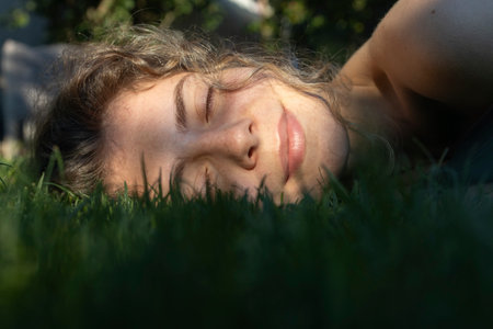 Close up of beautiful teenage girl, young adult woman lying on green grass in sunlight. Eyes closed,...