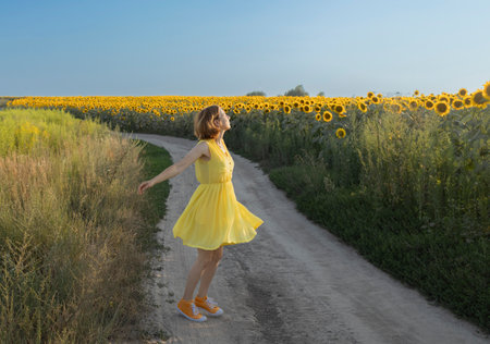 Beautiful happy woman in yellow dress dancing cheerfully near field of blooming sunflowers. Eyes closed enjoying nature outdoors. Cute girl relaxing walking in nature. summer, joy, freedomの写真素材