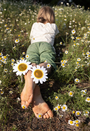 child lies on a meadow of wild daisies on a sunny day. On the heels of bare feet are funny glasses in the form of daisies. Happy childhood, be positive, playful mood, happy holidays. Hello, summerの写真素材