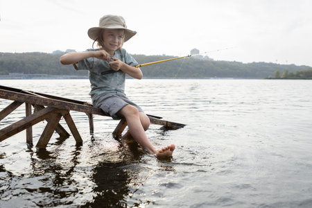 Funny boy 8 years old in a wide-brimmed hat with a fishing rod catches fish on a lake or river. Bare feet of a child in water. Healthy lifestyle, favorite hobby. Family vacation, summer entertainment.の写真素材