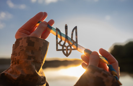Hands of a Ukrainian soldier holding a trident - a national symbol - with a yellow and blue ribbonの写真素材
