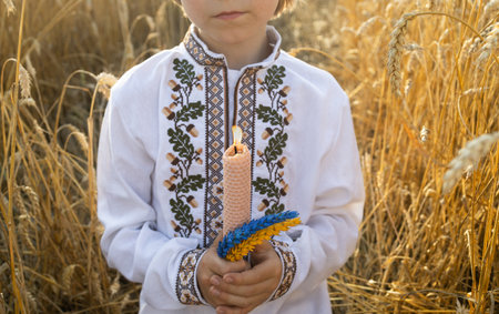 burning candle, two yellow and blue ears of wheat in colors of Ukrainian flag, in hands of boy in traditional embroidered shirt in wheat field. fire in memory of those who diedの写真素材