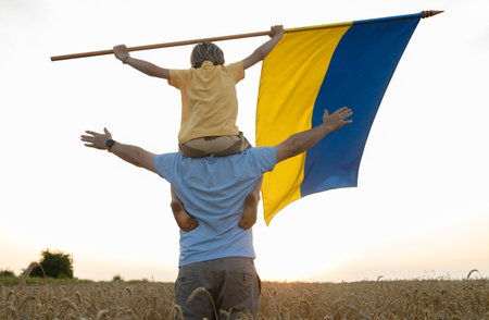Boy sitting on daddy's shoulders with a large satin flag of Ukraine in among wheat field at sunset. Patriotism education. Pride, freedom. Ukrainians against war. Symbol of country. Independence Dayの写真素材