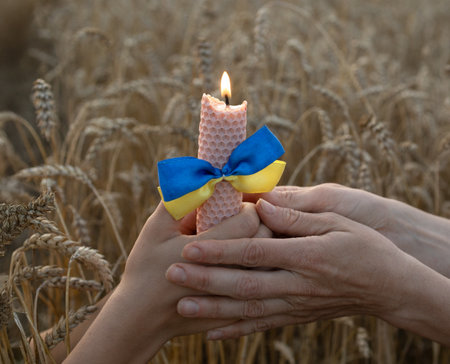Conceptual photo. A burning candle in the hands of an adult and a child among a wheat field. Ears of wheat and fire in memory of those who died during the tragic Holodomor in Ukraine in 1932-1933.の写真素材
