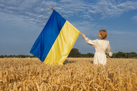 A woman holds a large Ukrainian flag standing in a golden sunlit wheat field against a blue sky. A national symbol of freedom and independence. Stop the war. Stand with Ukraine.の写真素材