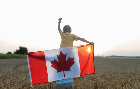 Canadian flag behind a boy sitting on the shoulders of an adult man standing in a wheat field at sunset. Happy Canada Day. Pride, freedom, patriotism. Traveling around the country. National symbol.の写真素材