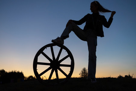 Interesting silhouette confident woman standing at sunset against sky, putting one foot on big wheel. Concept of readiness to meet new challenge, life energy, wheel of fortune. Search for inspirationの写真素材