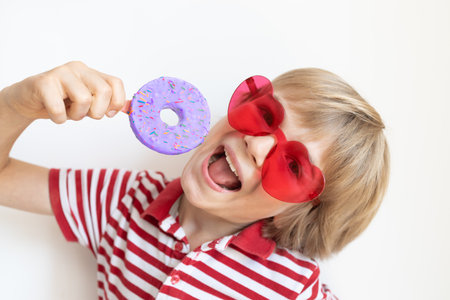 portrait of funny boy in red heart-shaped glasses having fun holding donut isolated on white background. Satisfied child preparing to bite tasty sweet dessert. Positive emotions, bright colors.の写真素材