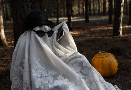 A child in a white blanket and black glasses sits under a tree in a gloomy forest and talks on the...