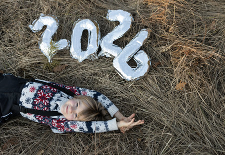 Silver numbers 2026, a happy child in a Christmas sweater lies on dry grass without snow. A symbol...