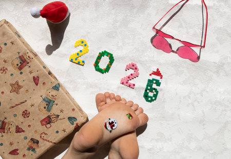 Multicolored numbers 2026, baby's bare feet, gift in wrapping paper and heart-shaped sunglasses on a...