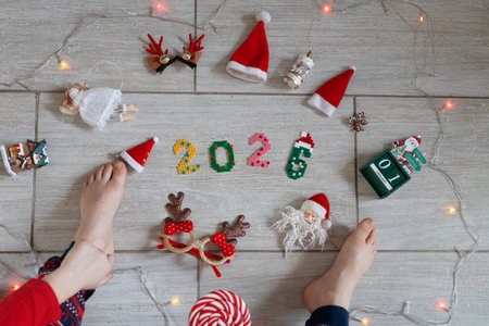 Multicolored numbers 2026, a child's bare feet, various Christmas decor and toys on a light floor. A...