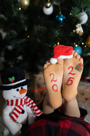 child's bare feet with numbers 2026 written on them, Santa hat on toes, toy snowman against backdrop of a Christmas tree. A festive atmosphere on New Year's Eve. A fun childhood, a humorous greetingの写真素材