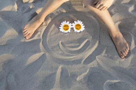 A barefoot child sits on the beach, drawing in the sand. A smiling sun made of sand and daisy-shaped sunglasses for eyes. Joy, relaxation, positivity, a happy childhood, sunbathingの写真素材