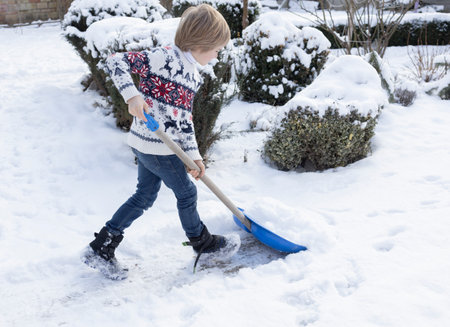 A boy in a winter sweater clears the yard of snow after a snowstorm, holding a large snow shovelの写真素材